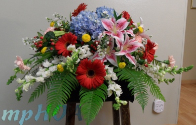 # MPCASK0011 — $275.00 — Blue Hydrangeas, Pink Oriental Lilies, Red Gerberas, White Snap Dragons, Pink Carnations, Red Hypericum, White Monte Casinos, Yellow Button Pom Poms. ~ Approximate size: 18″H x 36″W